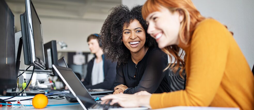 To smilende unge kvinner sitter ved skrivebordet på et kontor. Foran dem står det flere skjermer og en laptop.