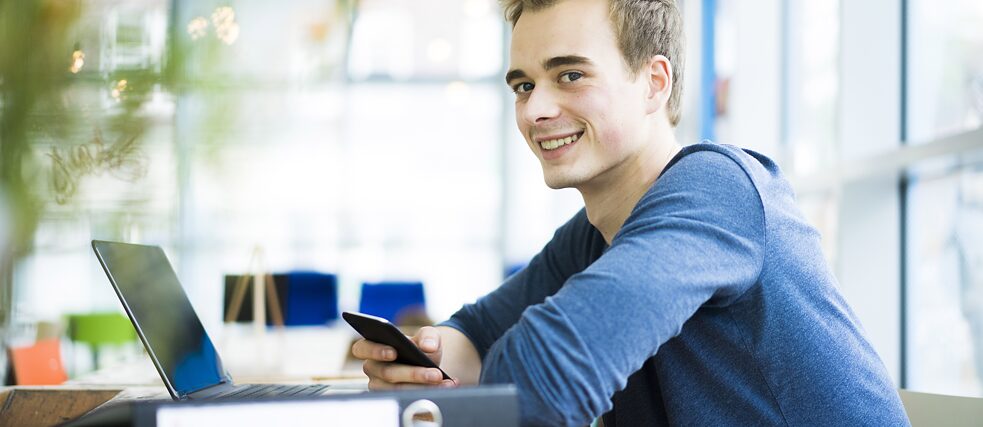 Ein junger Mann sitzt in einem Café. Vor ihm steht ein Laptop. Er hält ein Handy in der Hand und lächelt.