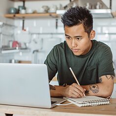 Ein junger Mann sitzt in der Küche vor einem Laptop und macht Notizen.