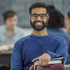Ein Student in einem Klassenraum blickt in die Kamera und lächelt. Im Hintergrund unscharf weitere Student*innen.