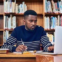 Ein Student arbeitet konzentriert am Laptop und macht Notizen. Im Hintergrund sind Bücherregale zu sehen.