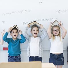 Primary school children are standing in front of a board, they are joking and holding books like a roof over their heads.