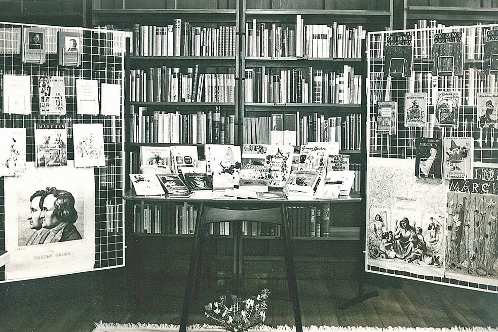 The picture shows a large book wall of the Grimms Fairy Tales exhibition, in front of which are two white metal grids on the left and right and a table in the middle. On the grids and on the table are various fairy tale books, on the grid also hang posters with further information about the authors and their works.