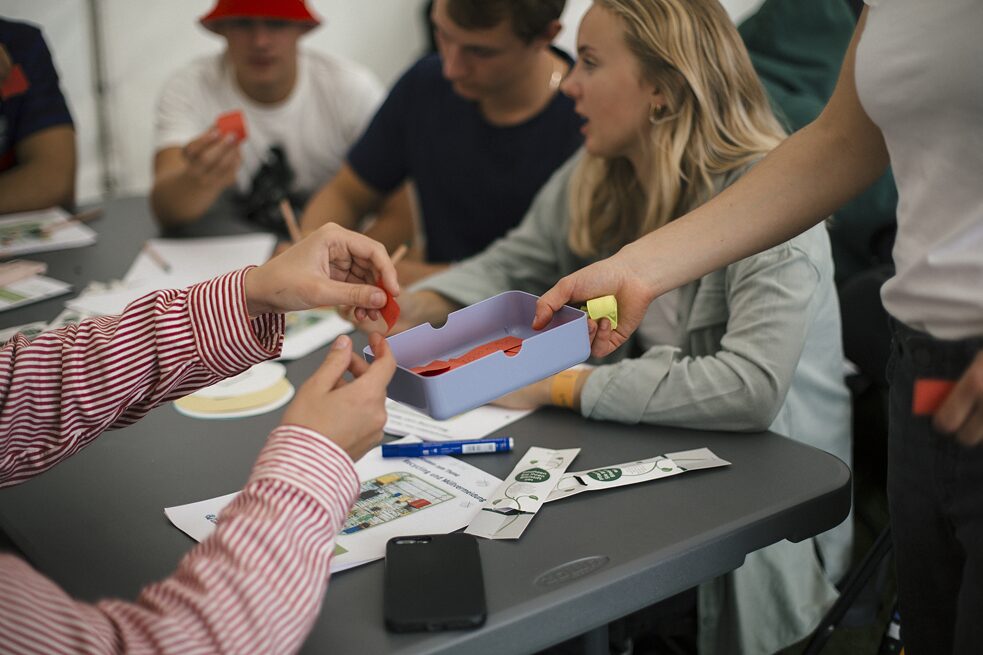 Fem elever sidder om et bord, hvorpå der ligger blyanter, opgaver og en mobiltelefon. Til højre i billedet uddeler en person små sedler fra en lille æske. 