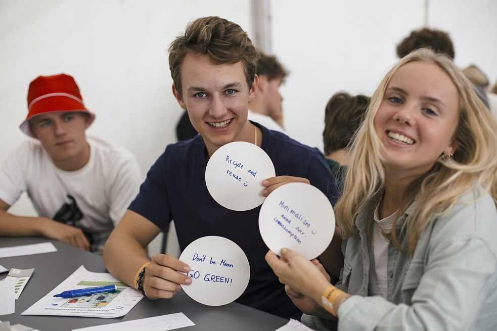 Tre elever sidder rundt om et bord. To af eleverne holder runde sedler op, hvorpå der står: "Don´'t be mean - go green", "recycle and reuse + smiley", "minimalism + smiley, avoid over-consumption".