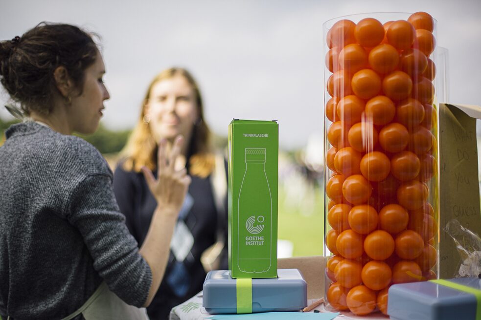 I billedets forgrund ses en gennemsigtig cylinder med orange plastikbolde i. Ved siden af står en madkasse og ovenpå en drikkedunk i en grøn karton. I baggrunden ses en person, der holder tre fingre op.  