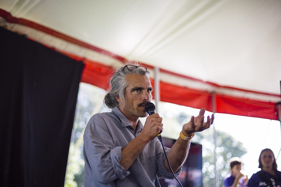 I midten af billedet ses foredragsholder, kunstner og aktivist Felix Becker. Han har sine briller siddende i panden og håret i en knold. I højre hånd holder han en mikrofon og venstre hånd rækker han ud foran sig.  
