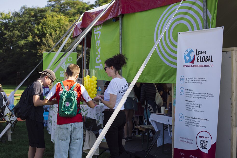 En person fra organisationen "Team Global" står sammen med to elever ved et højbord foran et telt. Eleverne står med ryggen til kameraet. På bordet står der en gennemsigtig cylinder med plastikbolde i. I baggrunden ses nogle træer. 