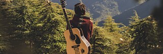 Ein Mann mit einer Gitarre auf dem Rücken steht in den Bergen, von hinten fotografiert