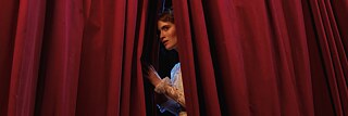 An actress on stage peering through an opening in the red curtains