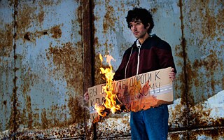 Bei einem seiner Einzelproteste hält Arshak Makichyan ein brennendes Schild aus Pappe auf dem steht „Ihr schafft die Wege zum Klimakollaps“.