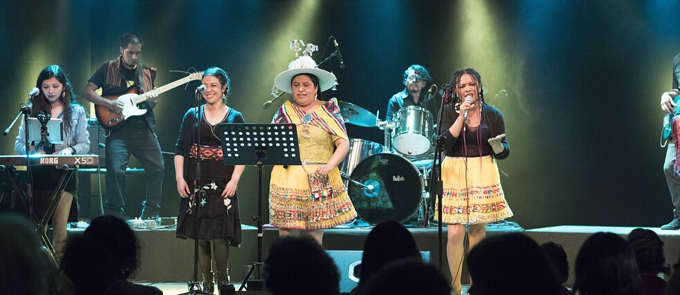 Drei Frauen treten auf Bühne auf und tragen traditionelle Kleidung. Sie werden von einer Band begleitet.