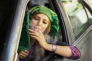 A woman sitting at the wheel of a car with her burqa flipped back and smoking a cigarette