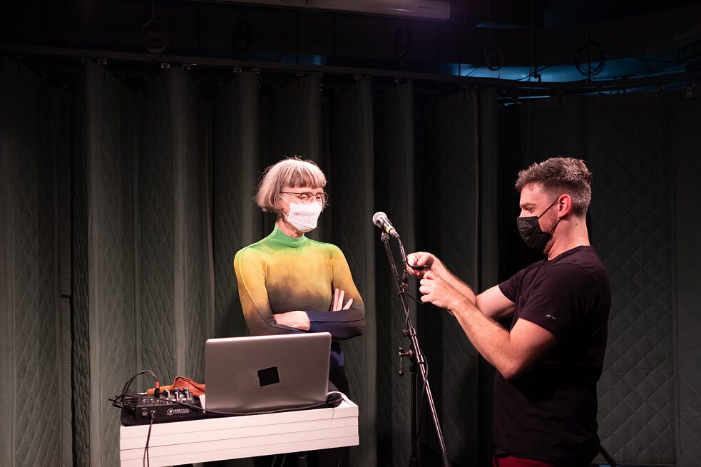 Foto von Ute Wassermann im Gespräch mit einem Mitglied des technischen Teams, während er ans Mikrofon arbeitet