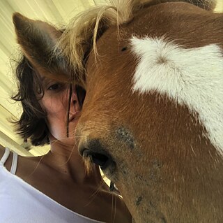 A girl in a white t-shirt and brown hair (Korallia Stergides) rests her head on the head of a brown horse.