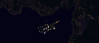 Auf dem Bild zu sehen ist die Insel Zypern als Satellitenaufnahme bei Nacht. Man kann zudem verschiedene gelbe Punkte und weiße Sterne auf der Insel erkennen, welche im dunklen Mittelmeerraum aufleuchten.