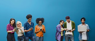 7 mladih ljudi stoje jedno pored drugog i zajedno gledaju u mobitele ili tablete