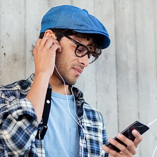 A young man in a blue beret and checked shirt holds a smartphone in his hand and wears in-ear headphones.