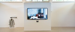 A TV screen with headphones and an artwork with a hanging raincoat are placed on a wall.