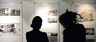 Ein Mann und eine Frau stehen mit dem Rücken zur Kamera und blicken auf drei Ausstellungsbanner, die an einer Wand hängen.