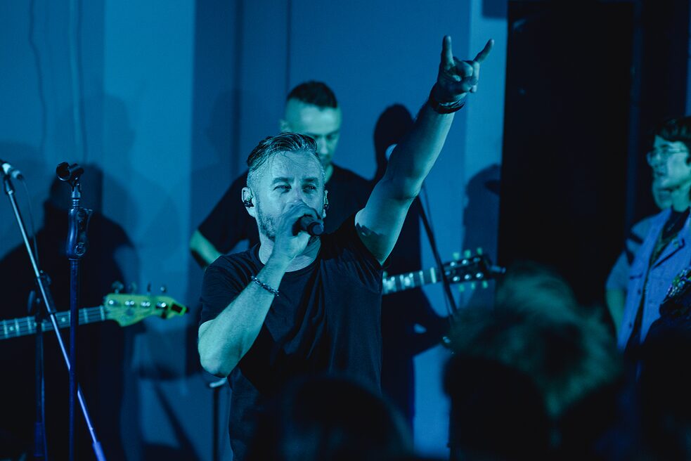 Serhij Zhadan zeigt mit seiner linken Hand die "Metalhand" (englisch "Devil horns"). Hinter ihm ist seine Band "Zhadan I Sobaki" zu sehen. Alle sind schwarz gekleidet. 