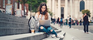 Eine Schülerin sitzt in der Stadt auf einer Treppe und arbeitet mit dem Laptop. Neben ihr steht ein Coffee to Go, liegen Arbeitsunterlagen und ein Rucksack.