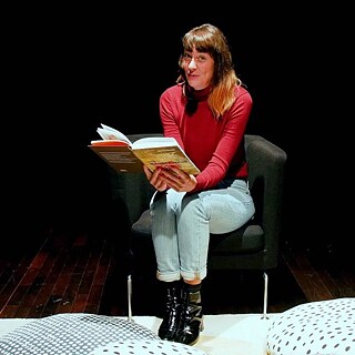 Deborah Weber lit un livre assise sur un fauteuil. Des coussins sont posés sur le sol devant elle.