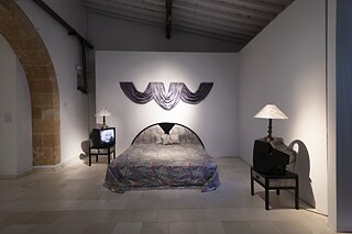 An interior design installation consisting of a double bed and two small tables on top of which are old-fashioned TVs and a table lamp.