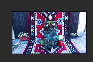 Film still on a big screen showing a woman sitting on a colourful carpet holding half a watermelon. Next to her are two more watermelons.