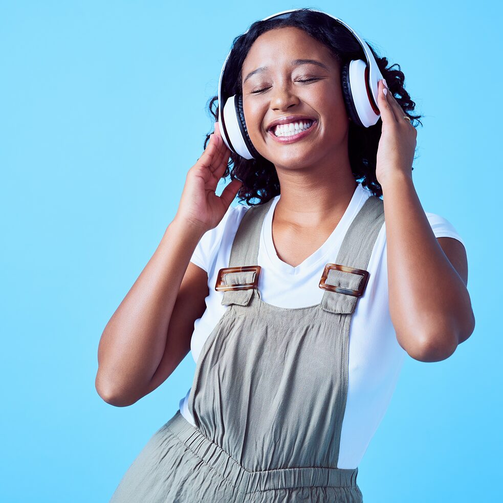 Eine lachende junge Frau hört mit geschlossenen Augen und weißen Kopfhörern Podcast vor blauem Hintergrund.