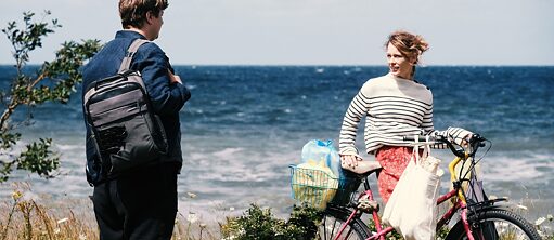 Filmscene fra Christian Petzolds film "Red Sky": Nadja, spillet af Paula Beer, står ved kysten med en cykel og havet i baggrunden. Hun smiler til Leon, spillet af Thomas Schubert, som står med ryggen til kameraet og vender sig mod Nadja.