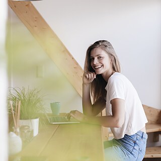 Eine junge Frau sitzt am Tisch vor einem Laptop und lächelt in die Kamera.