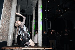 A woman stretches her arms into the air in a dance-like manner. She kneels on a wooden bench surrounded by stone pillars. 