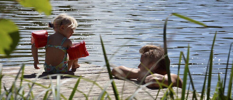 Ein Kind sitzt mit Badeanzug und Schwimmflügeln zusammen mit dem Vater an einem See.
