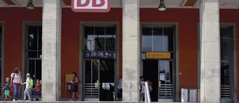 Ansicht auf ein Bahnhofsgebäude, oben ist ein abgeschnittenes Deutsche Bahn-Logo zu sehen, außerdem Wegweiser zu den Gleisen.