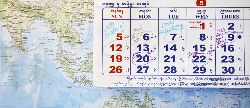 Vor einer Weltkarte hängt ein Wandkalender auf Burmesisch mit deutschen Notizen.