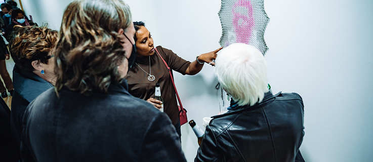 Drei Frauen betrachten ein Kunstwerk an der Wand, eine Frau zeigt darauf und erklärt etwas