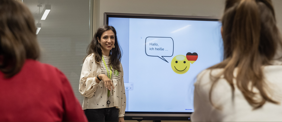 A woman smiles and stands at the whiteboard. On the board you can read the sentence "Hallo, ich heiße..." We see students from behind.