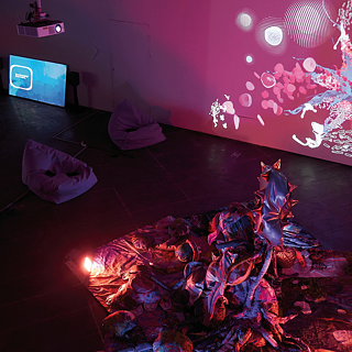 Ein künstlerisch inszeniertes Bild zeigt einen Raum mit einer Projektion abstrakter, leuchtender Muster an der Wand, ein Bündel schimmernder Stoffe auf dem Boden.