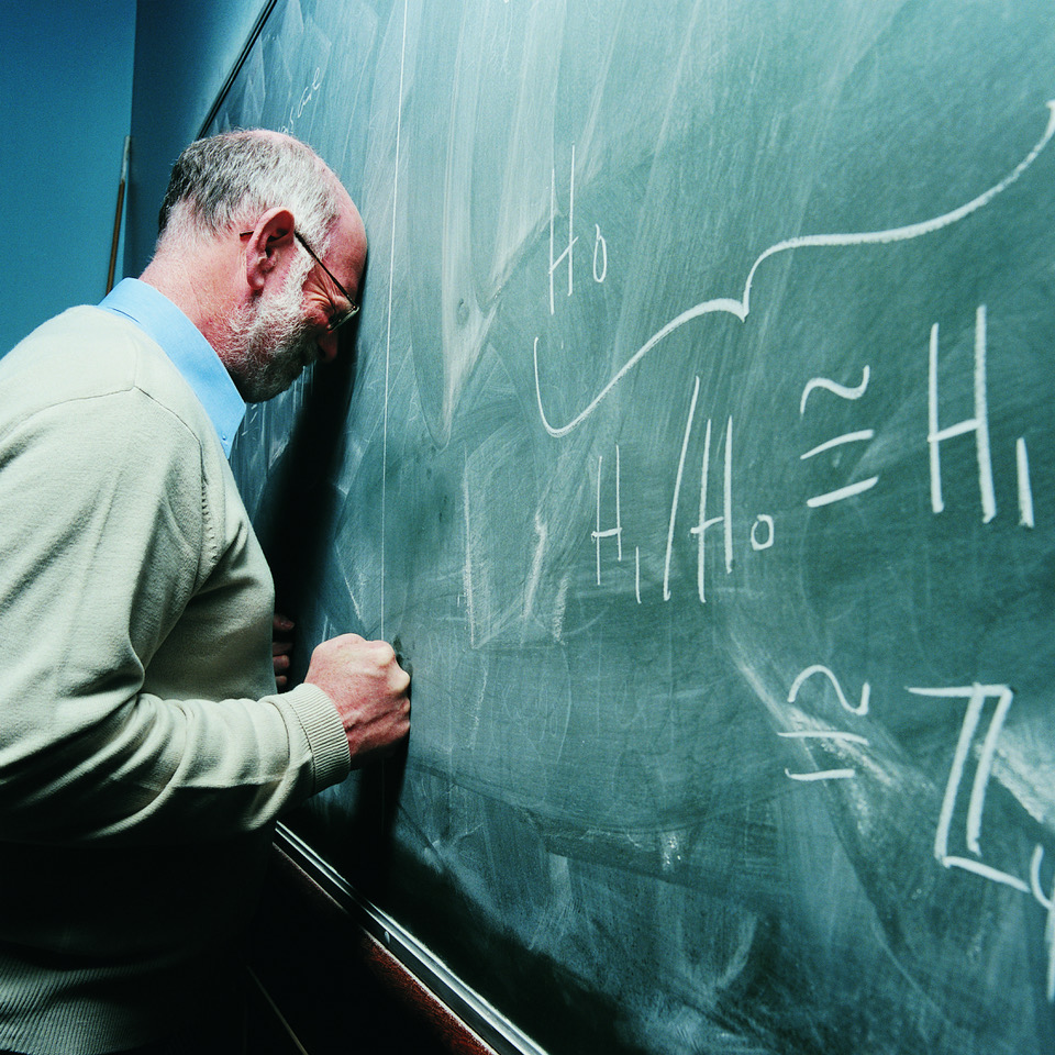 An elderly teacher stands in front of a large, written board. Frustrated, he leans his head and clenched fist against the blackboard.