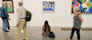 Woman sits in a gallery and observes a painting as others are passing by