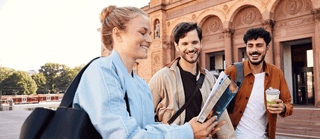Drei junge Erwachsene, eine Frau und zwei Männer, stehen fröhlich vor einem rötlichen historischen Gebäude. Die Frau im Vordergrund hält eine Broschüre in der Hand und zeigt ihren beiden männlichen Begleitern etwas darauf. Alle drei wirken entspannt, neugierig und gut gelaunt.