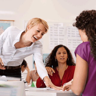 Kaks täiskasvanud õppijat istuvad ümber laua. Õpetaja kummardab üle laua, et ühele õppijale midagi näidata. Kõik on huvitatud ja naeratavad. Taustal istuvad veel kaks õpilast, seinte peal ripuvad kaardid ja õppeplakatid.