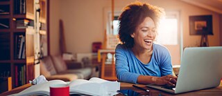 Eine junge Studentin arbeitet zu Hause an ihrem Laptop. Sie sieht auf den Bildschirm und lacht.