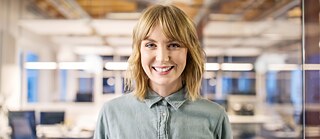 A young woman is smiling at the camera. In the background is a large office.