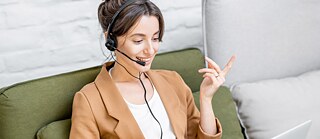 A young woman is sitting on a sofa. She is taking part in an online meeting and wearing a headset.