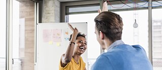 Zwei fröhliche junge Menschen klatschen die Hände gegeneinander und lachen. Im Hintergrund steht eine Whiteboard.