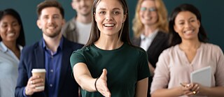 Eine Gruppe Mitarbeiter*innen eines Unternehmens, eine Frau streckt die Hand zum Handschlag entgegen.