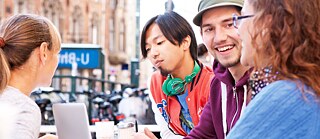 To unge mænd og en kvinde sidder udendørs på en café og arbejder på et projekt.