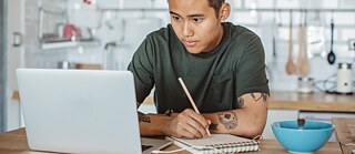 Ein junger Mann sitzt in der Küche vor einem Laptop und macht Notizen.